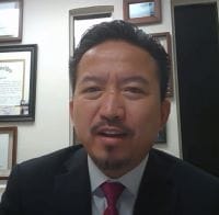 A man in a suit and red tie is speaking to the camera about civil engineering. He is in an office with framed certificates on the wall behind him, emphasizing the benefits of outsourcing key projects for efficiency.