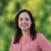 A person with long dark hair and a pink blouse smiles confidently, reminiscent of someone who's just mastered civil engineering outsourcing, standing in front of a blurred green background.