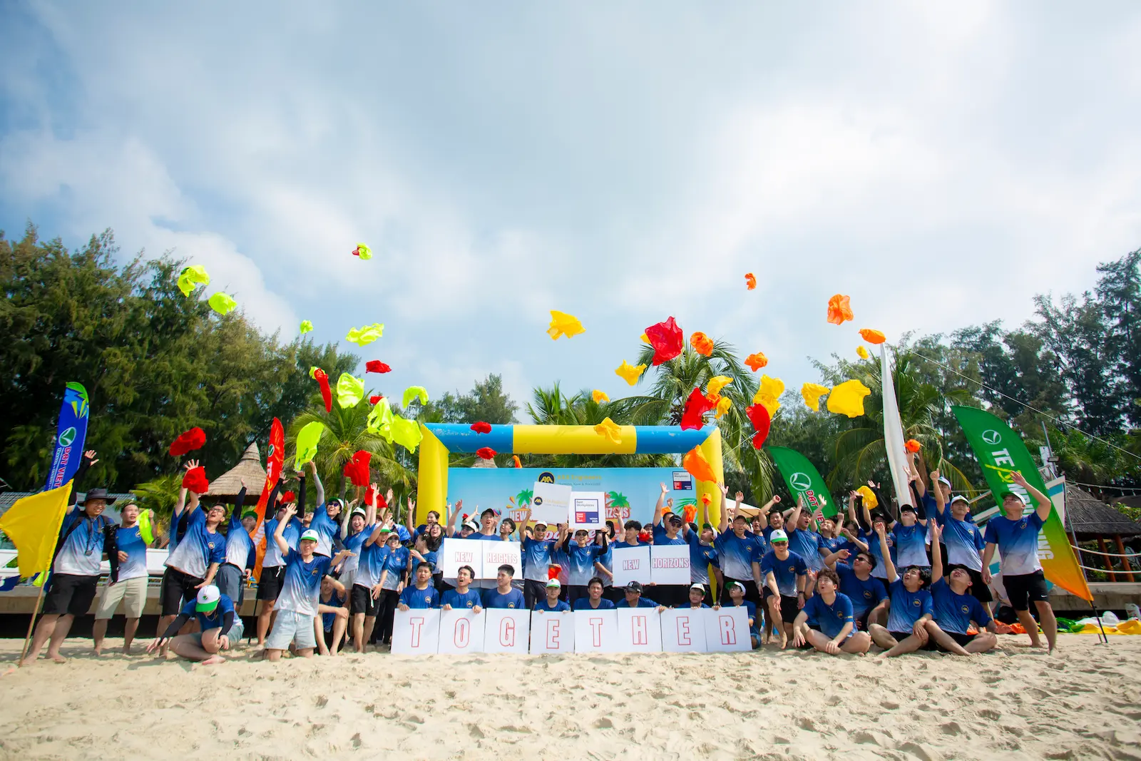 Group in blue shirts on a beach toss colorful fabric, with banners and TOGETHER sign behind.