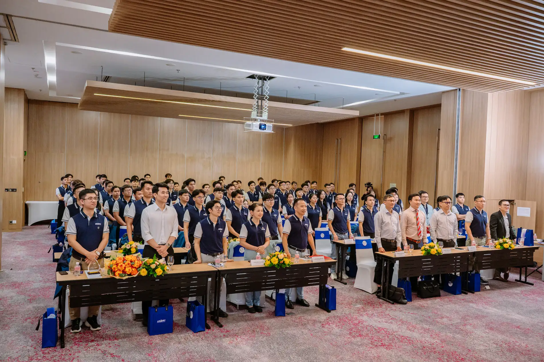 People in navy shirts stand behind tables with blue gift bags at an AXANH recruitment event.