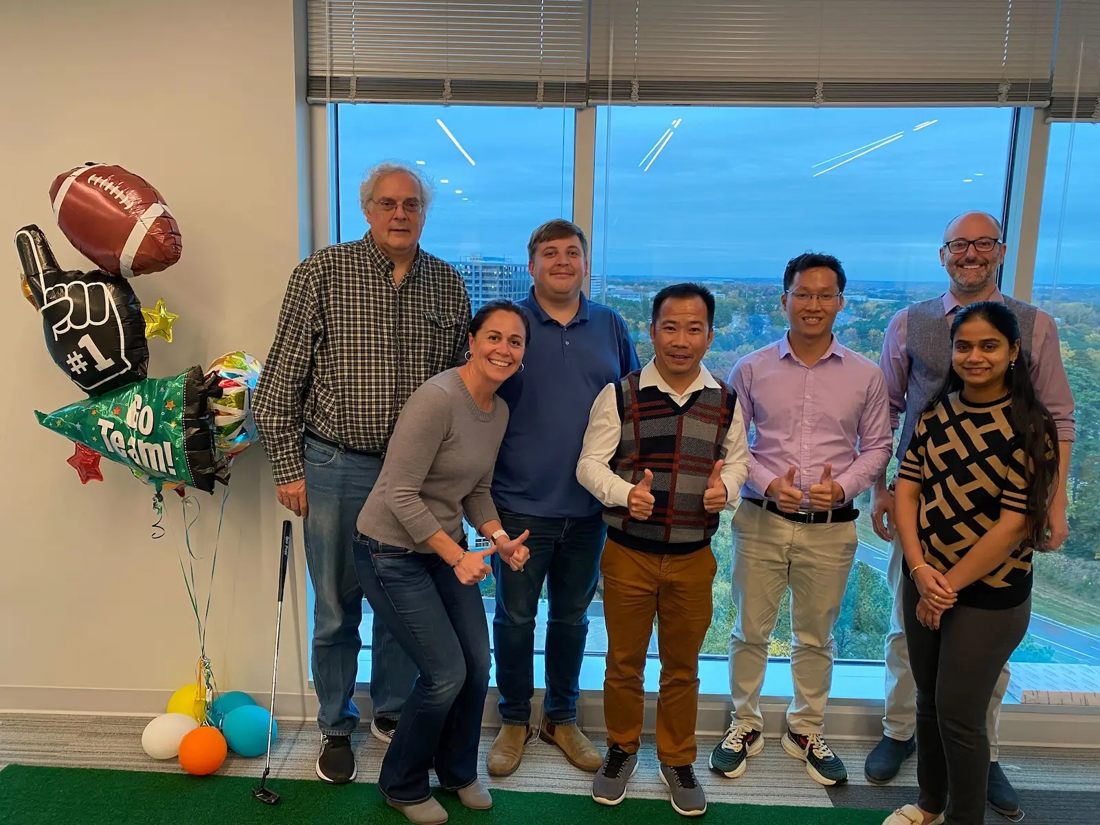 Seven people smile by a window with football balloons and a city view; putting mat visible.
