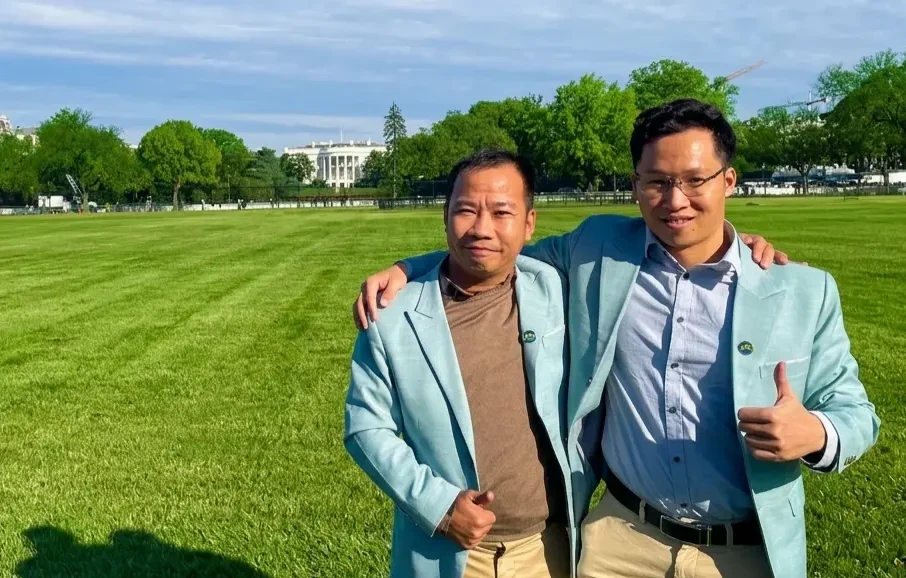Two men in light blue blazers stand smiling together on a grassy lawn, arms around each other.