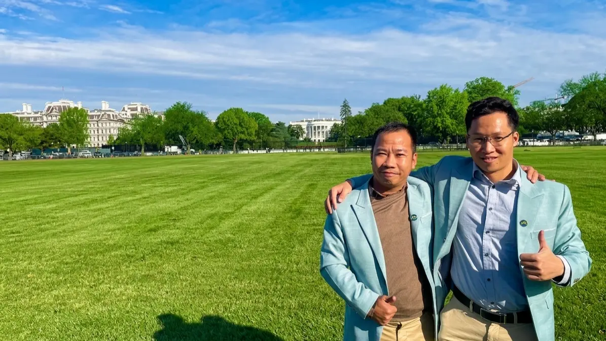 Two men in light blue blazers stand smiling together on a grassy lawn, arms around each other.