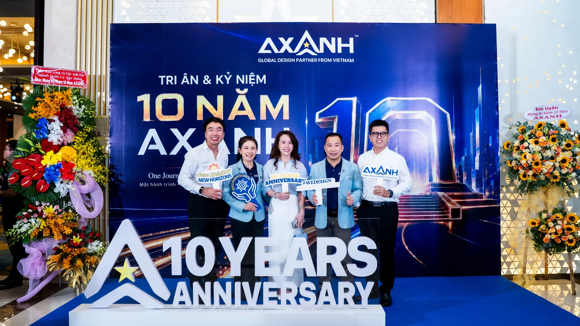 Five people smile at the AXANH 10th anniversary with themed signs and backdrop.