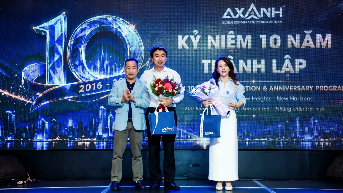 Three people on stage with flowers and gift bags at AXANH 10th anniversary.