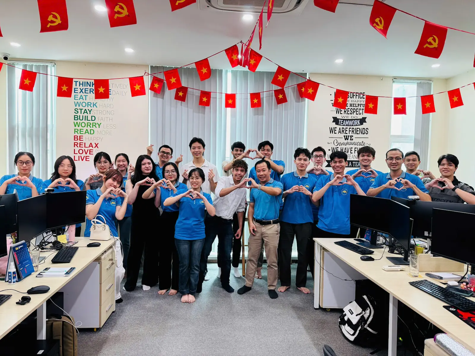 Office group posing, many making heart signs, Vietnam flags overhead, smiling.