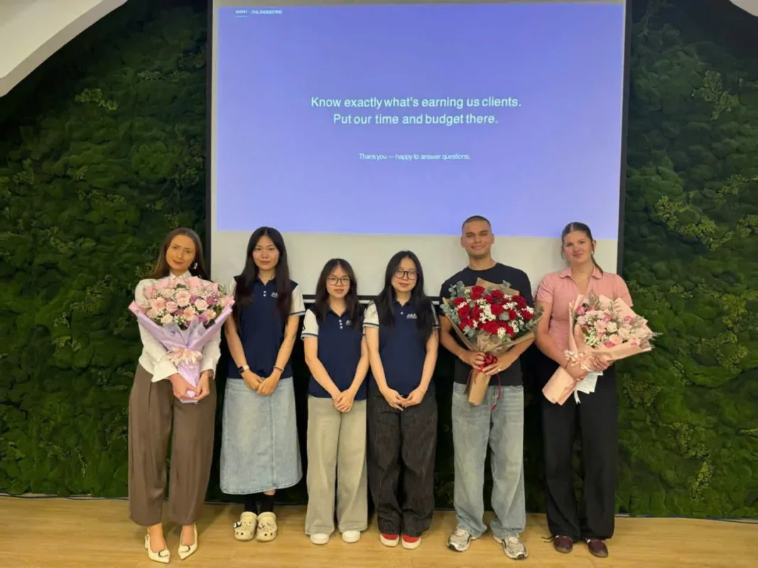 Six people indoors with bouquets, smiling before a screen and green plants.