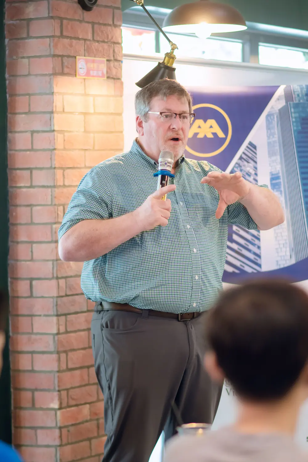 Man in glasses and green checked shirt speaks, gesturing, at yellow logo banner.