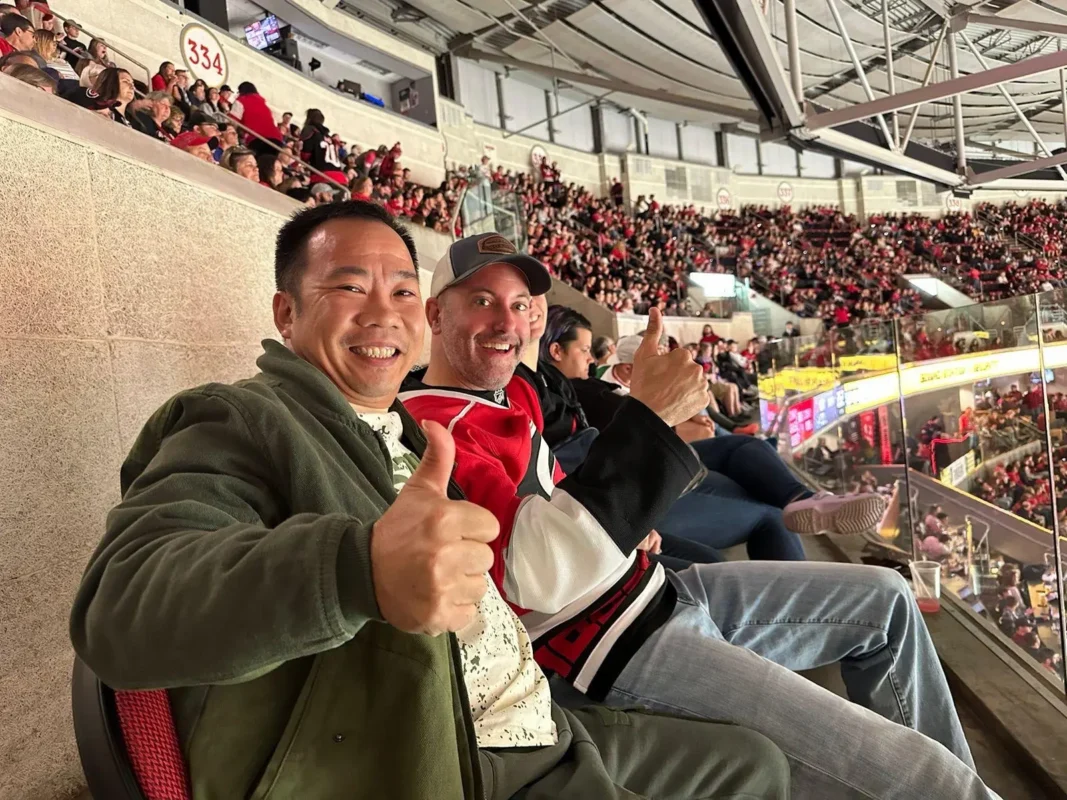Two men in stadium seats smile and give thumbs up at a hockey game.