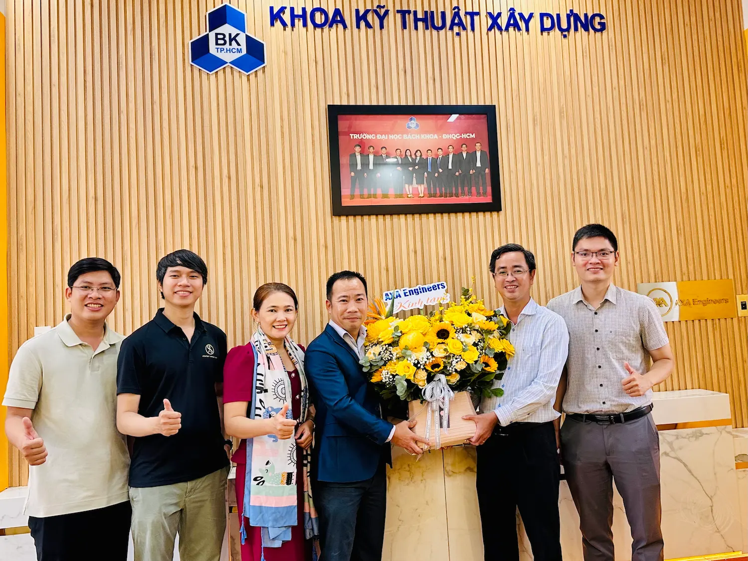 Six people indoors smiling, holding yellow flowers; four give thumbs-up. Vietnamese wall text visible.