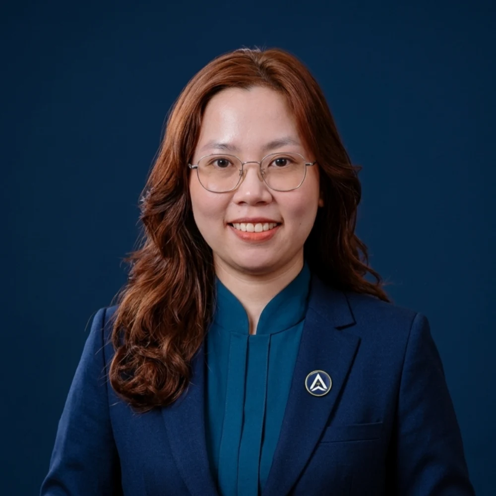 Woman with wavy brown hair, glasses, blue jacket, smiles on dark background.