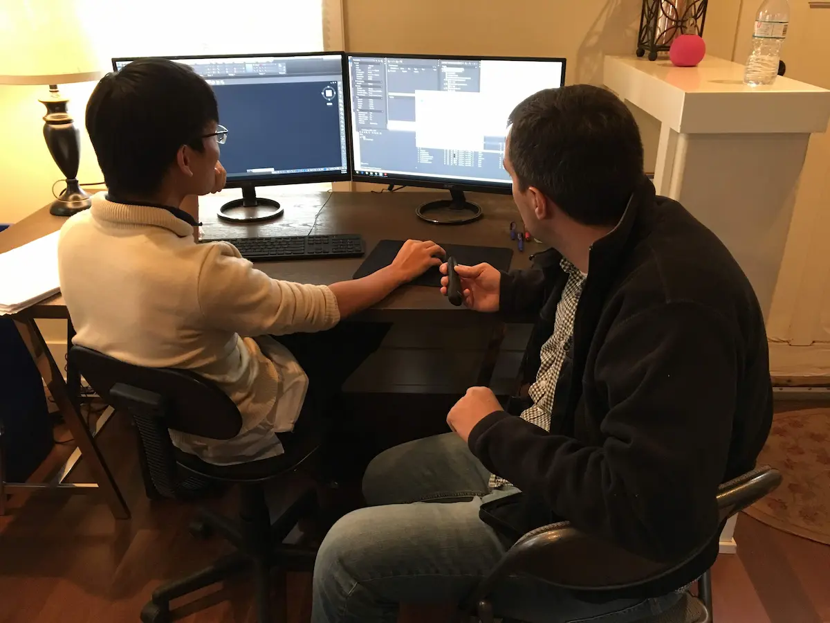 Two people at a desk; one types, the other points at code on screen.