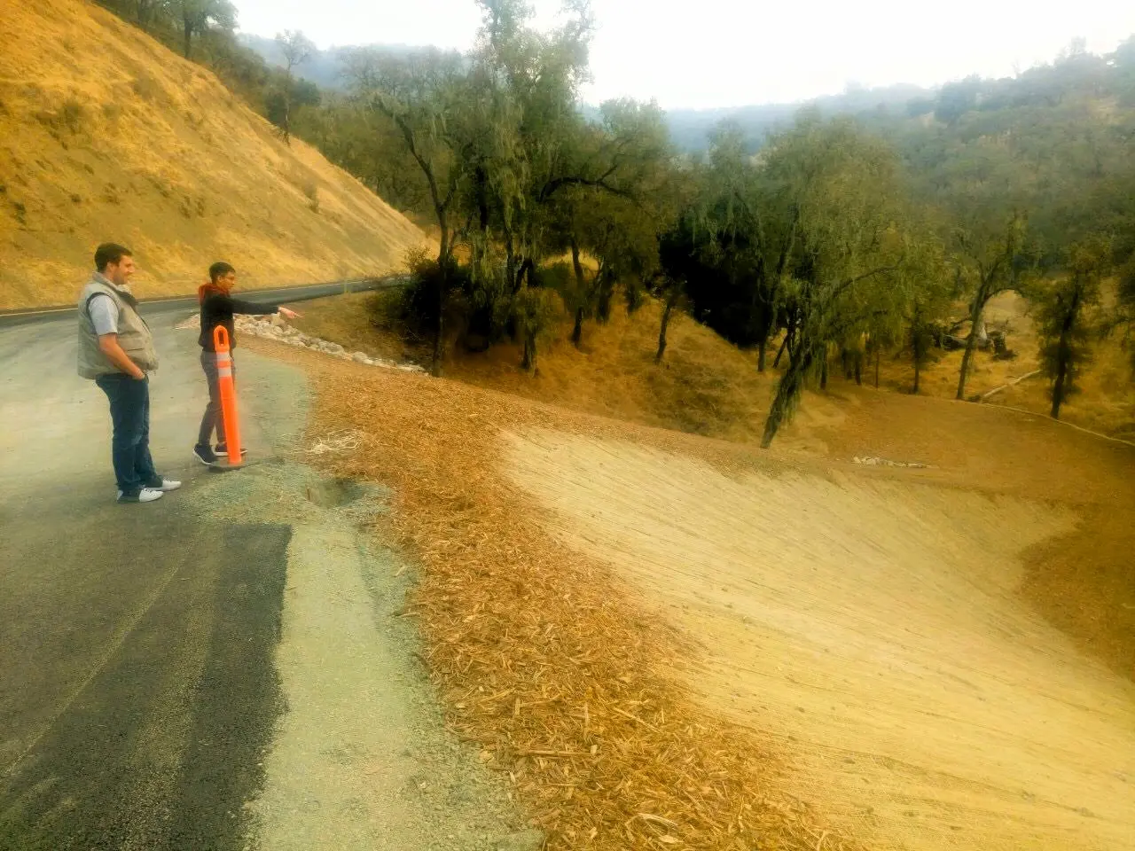 Two people stand by a road and orange post, looking at dry hills.