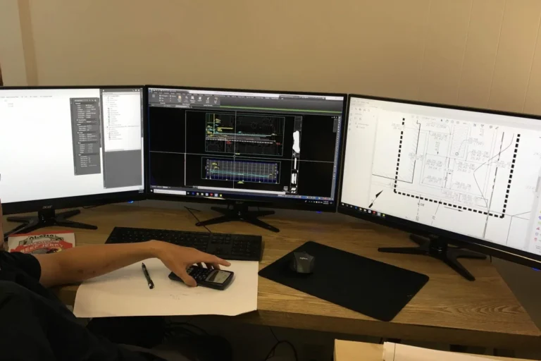 Person at desk with three monitors, papers, calculator, pen, and mouse.