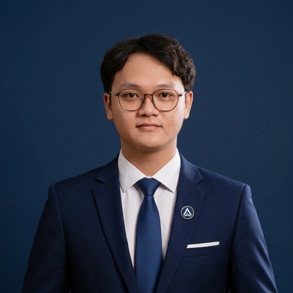 Young man in a navy suit and glasses stands against a dark blue background.