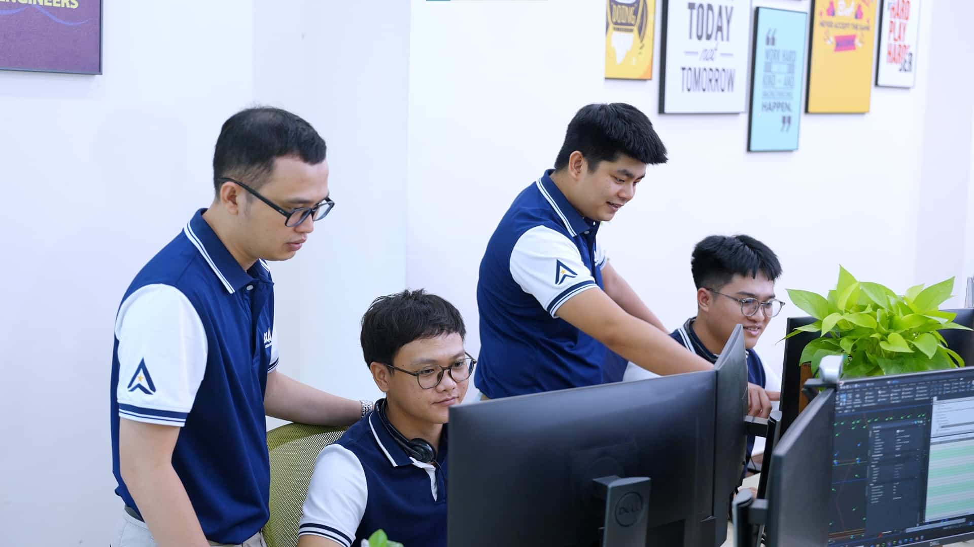Four men in navy and white shirts work together at computers in an office.