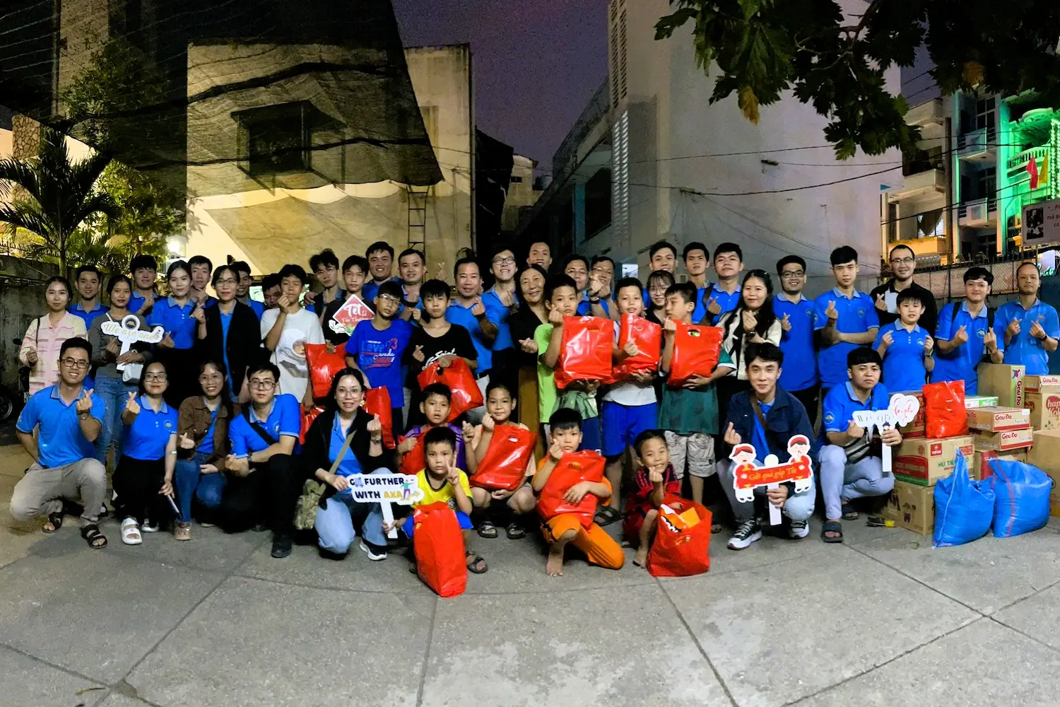 Group of adults and children smiling outdoors at night with gift bags and signs.