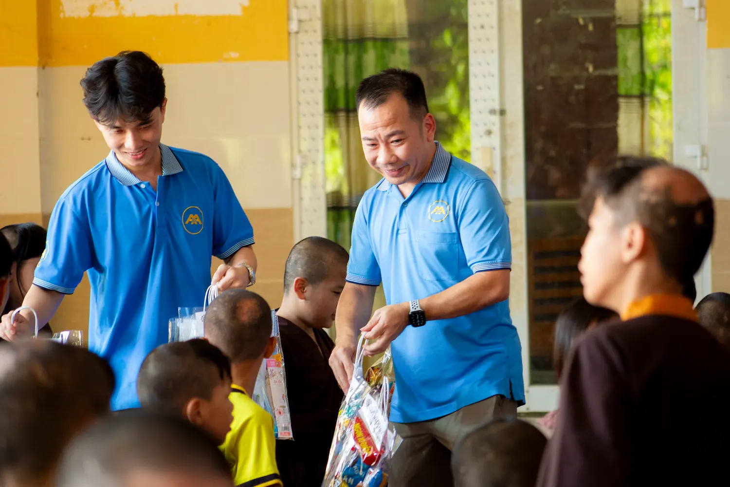 Two men in blue shirts give gift bags to seated children in a yellow room.