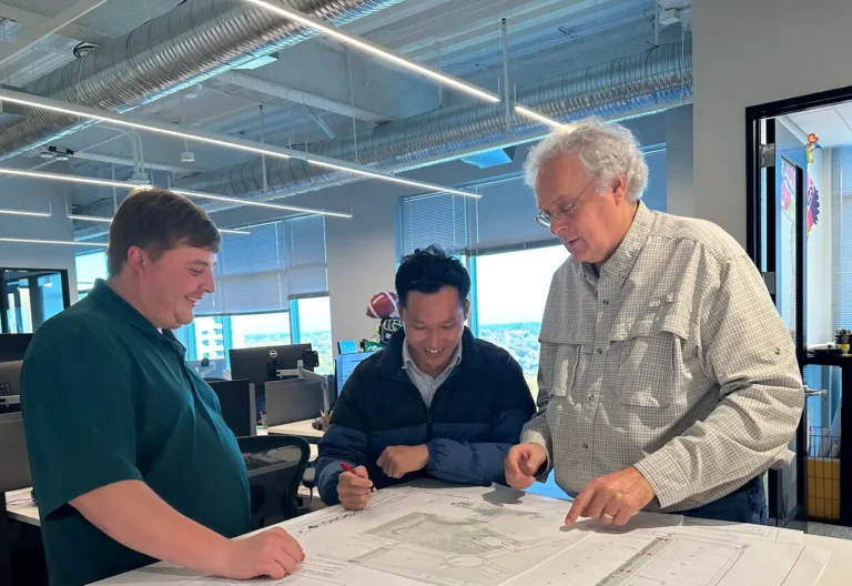 Three men discuss blueprints at a table in a bright, modern office.