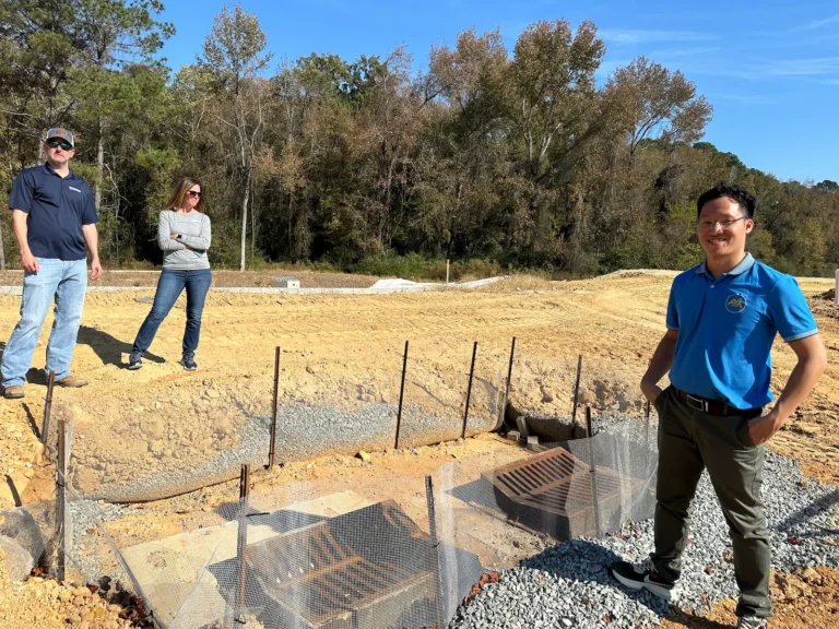 Three people by a construction site with drain grates, fencing, and nearby trees.
