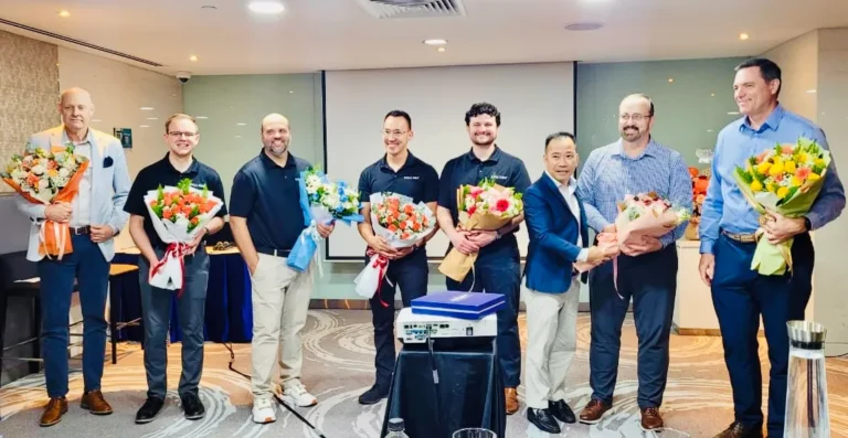 Eight men indoors, seven holding bouquets and smiling, in business casual attire.