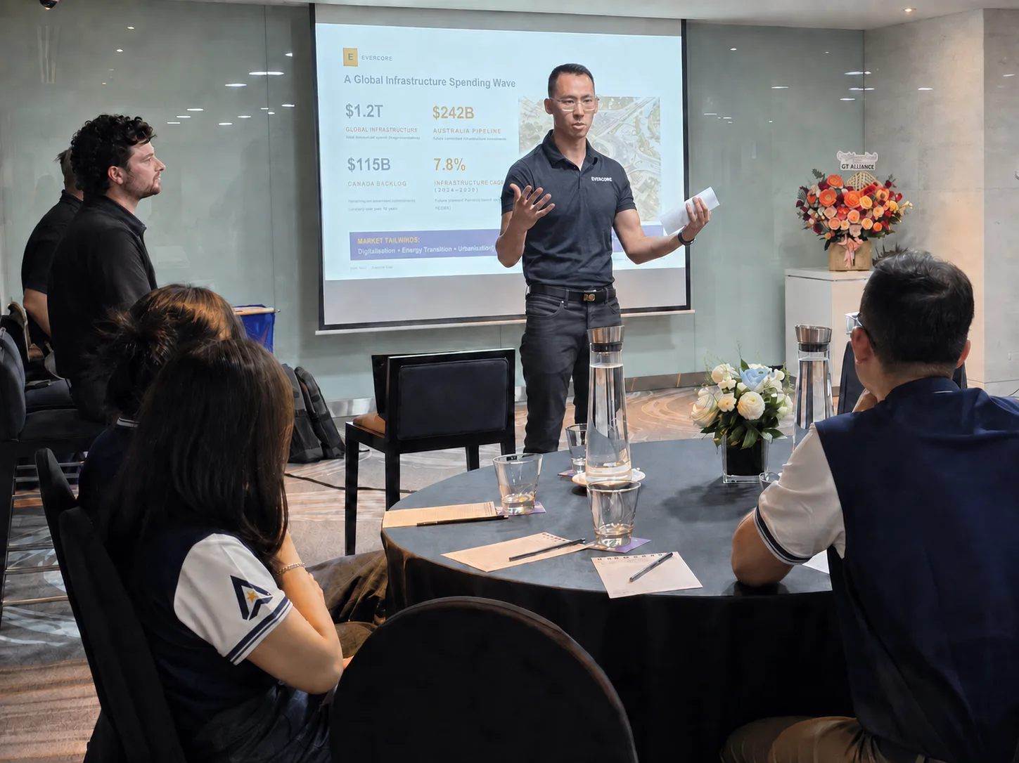 Man presenting to seated group; projector displays global infrastructure data; tables set.