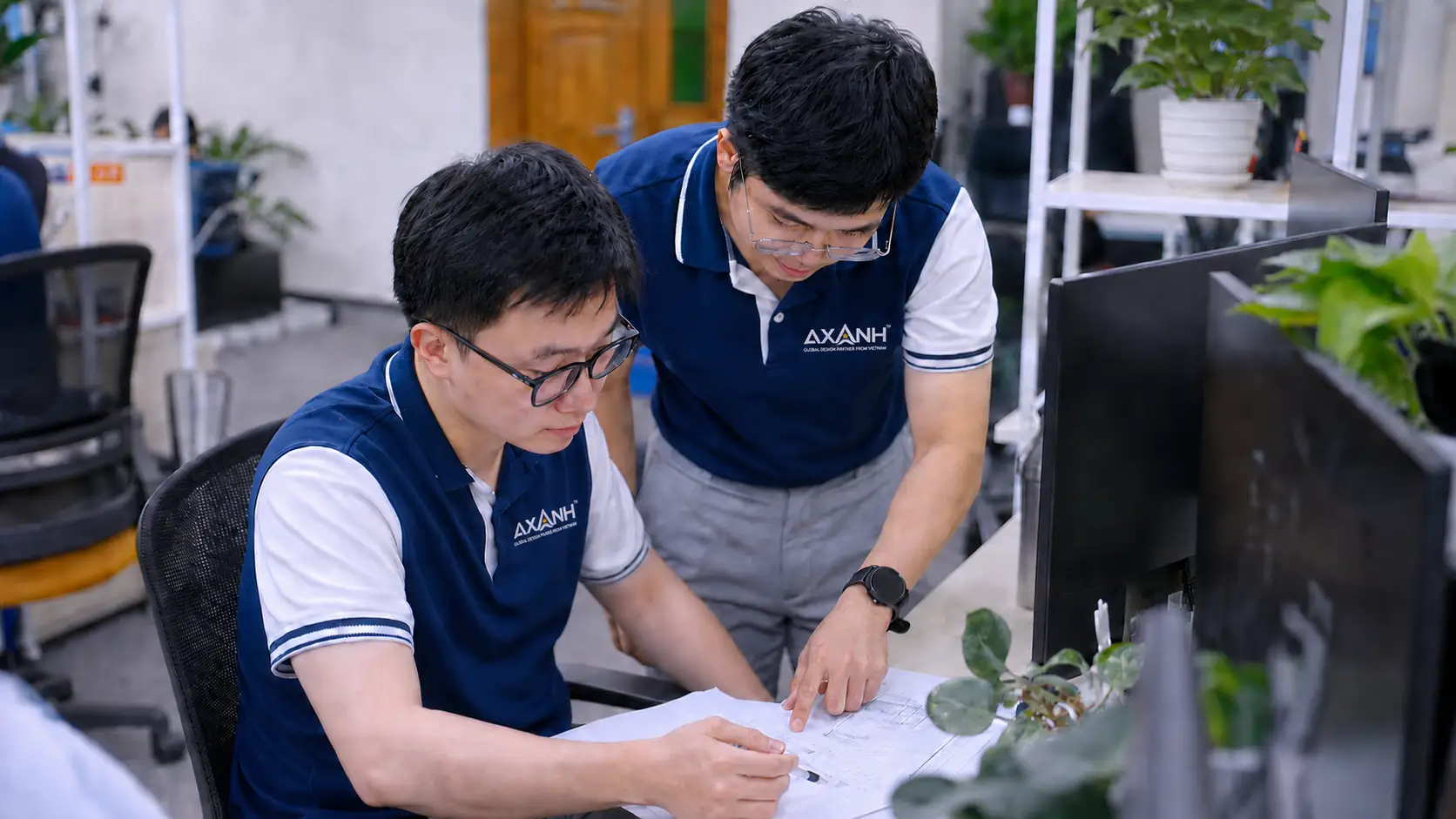 Two men in blue and white shirts discuss documents at an office desk.
