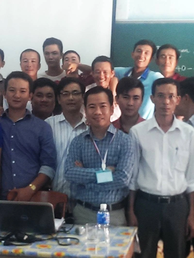 Men stand and sit in a classroom; chalkboard, laptop, and water bottle visible.